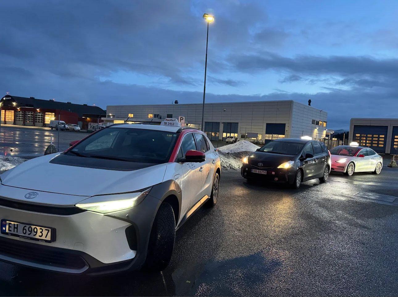 En hvit taxi og flere biler står parkert på en gate i skumringen, med bygninger i bakgrunnen.
