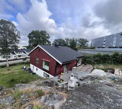 Rødt hus med svart tak, plassert på en steinete hage med trær og moderne bygninger i bakgrunnen.