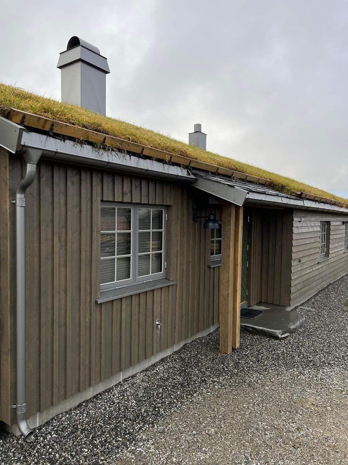 Hus med torvtak og trevegg, beliggende på grusvei under en skyet himmel.