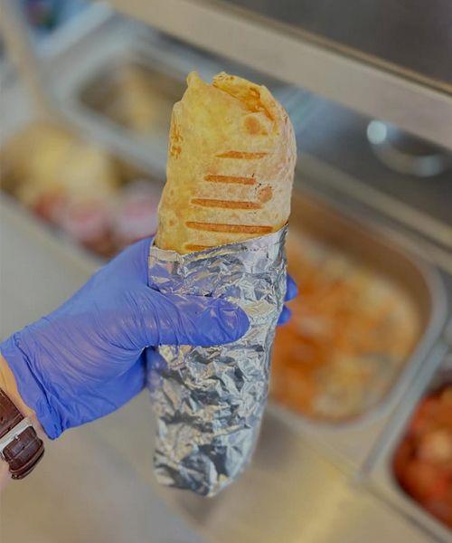 En hånd som holder en innpakket burrito med grillet overflate, med matvarer i bakgrunnen.