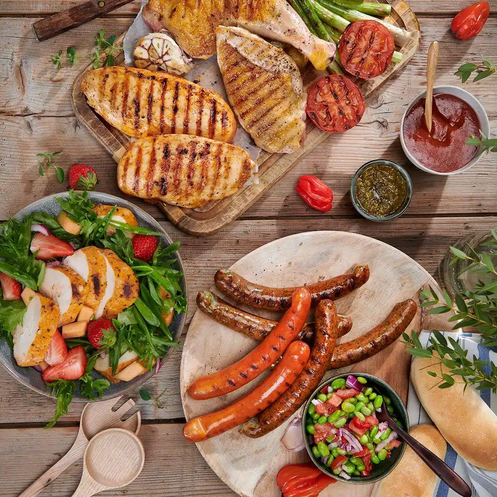Grillmat med kylling, pølser, grønnsaker og salat på et trebord, perfekt for en sommerfest.