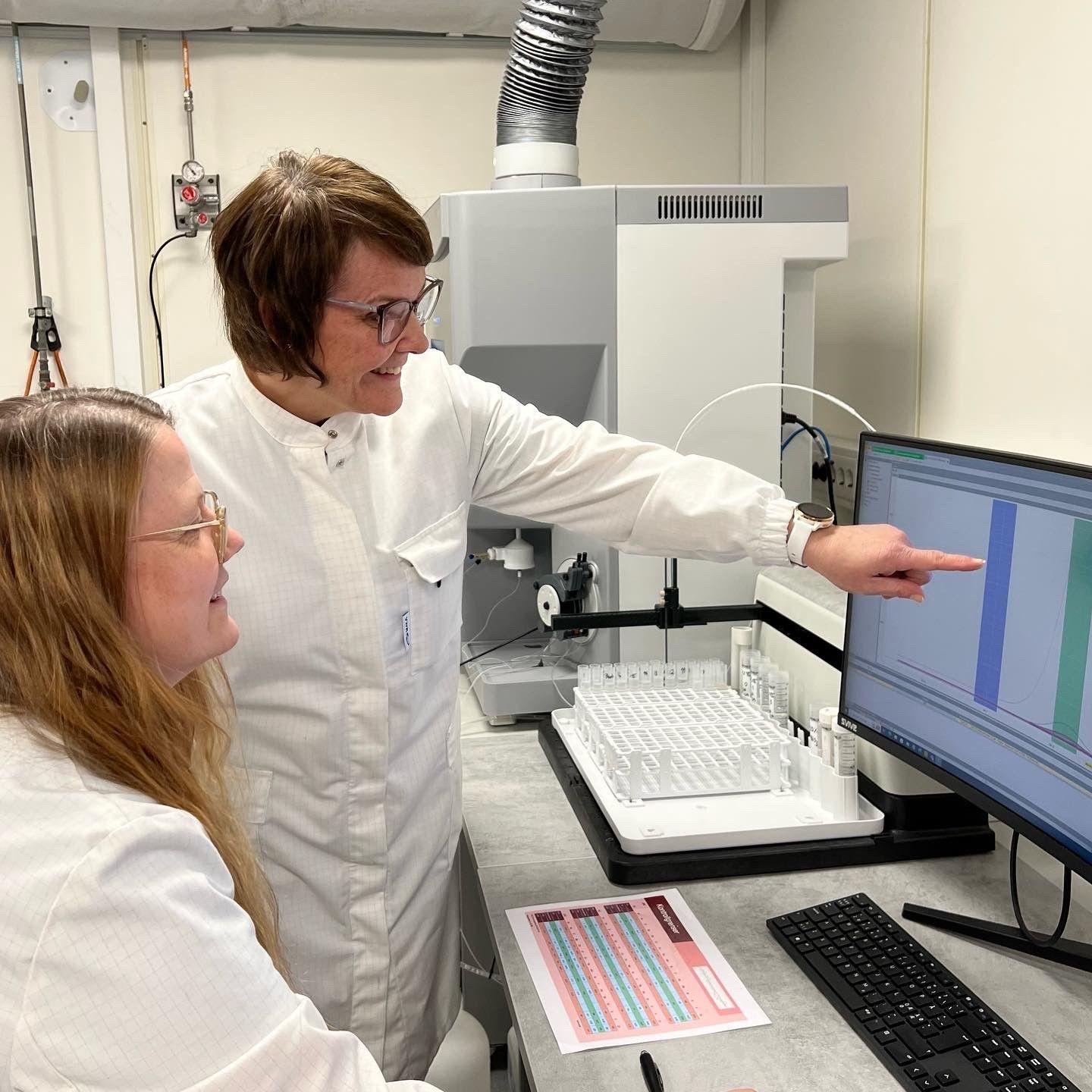 To kvinner i labfrakker ser på en dataskjerm med analyseresultater i et laboratorium.