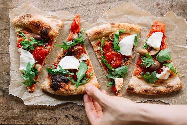 Hånd som tar en skive pizza med tomatsaus, mozzarella, ruccola og oliven på et trebord.