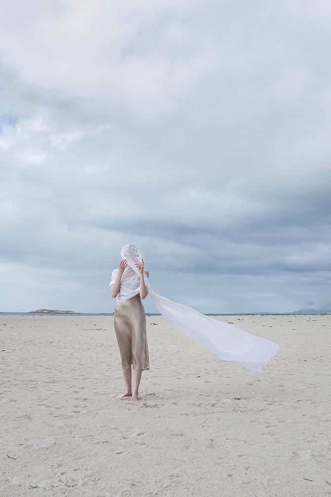 Kvinne med hvit sjal som står på stranden under en skyet himmel. Sanden er lys og landskapet er åpent.
