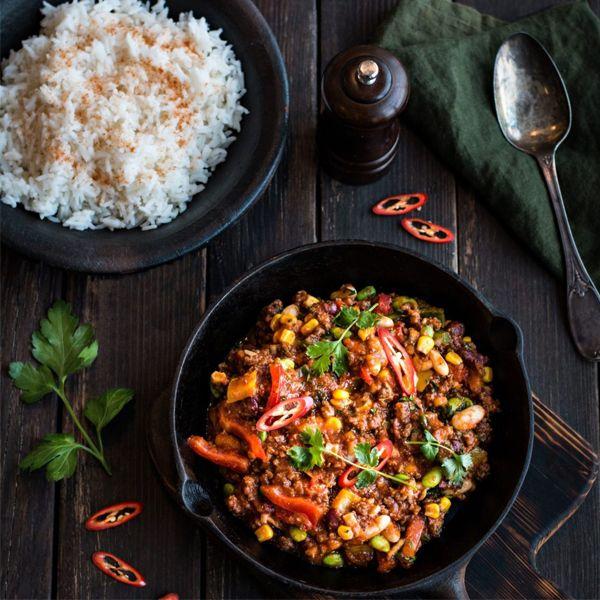 Krydret kjøttdeiggryte med bønner og paprika, servert med hvit ris og chiliskiver på en mørk treoverflate.