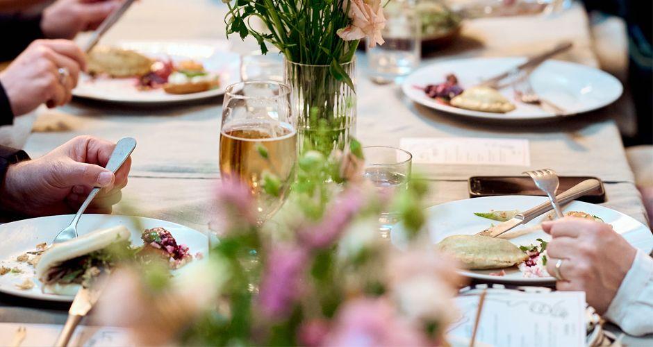 Borddekk med tallerkener, bestikk og blomster, der folk nyter middag sammen.
