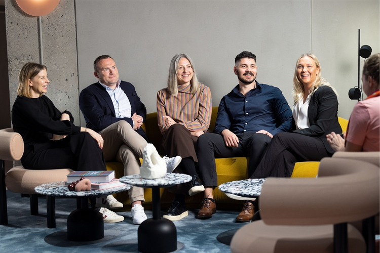 En gruppe på fem personer sitter sammen i en moderne stue med sofaer og bord. De smiler og ser glade ut.
