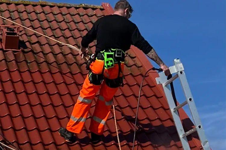 En håndverker i oransje arbeidsklær står på et tak og bruker en stige under blå himmel.