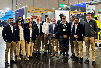 Gruppe av mennesker i formelle klær som poserer på en messe med utstillingsstander i bakgrunnen.
