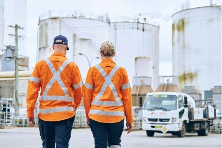 To personer i oransje arbeidsklær med refleksstriper går mot en lastebil ved industrielle tanker.