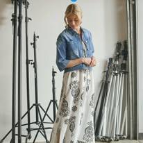 Kvinne i en lang, mønstret kjole med denimjakke, står foran fotostativ i studio.