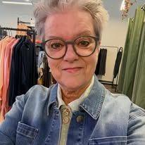 En eldre kvinne med grått hår og briller smiler i en klesbutikk, iført en denimjakke.
