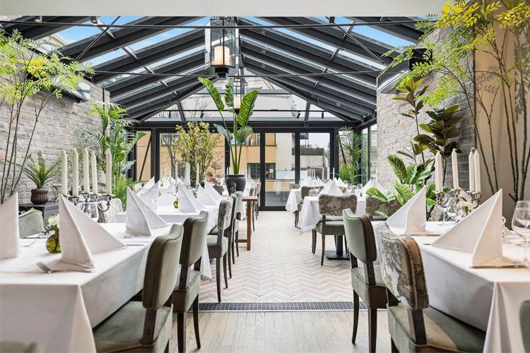 Lyst og moderne restaurantinteriør med hvite duker, planter og borddekorasjoner i et glasshus.