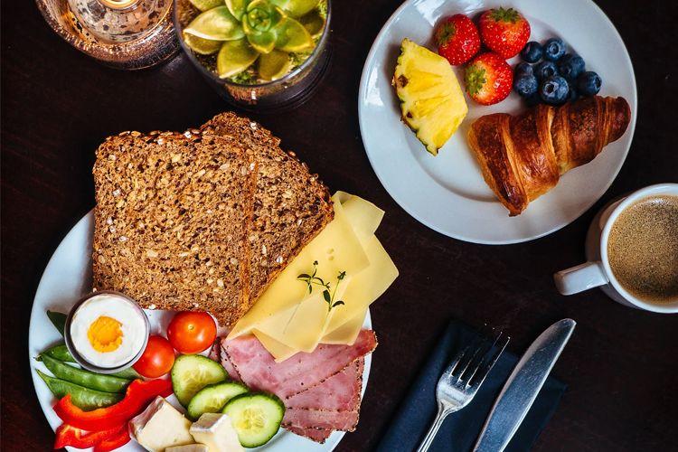 Frokosttallerken med grovt brød, ost, skinke, grønnsaker, egg, frukt og en croissant, servert med kaffe.
