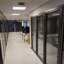 Moderne kontorlandskap med glassvegger, skrivebord og stoler i en lys korridor.
