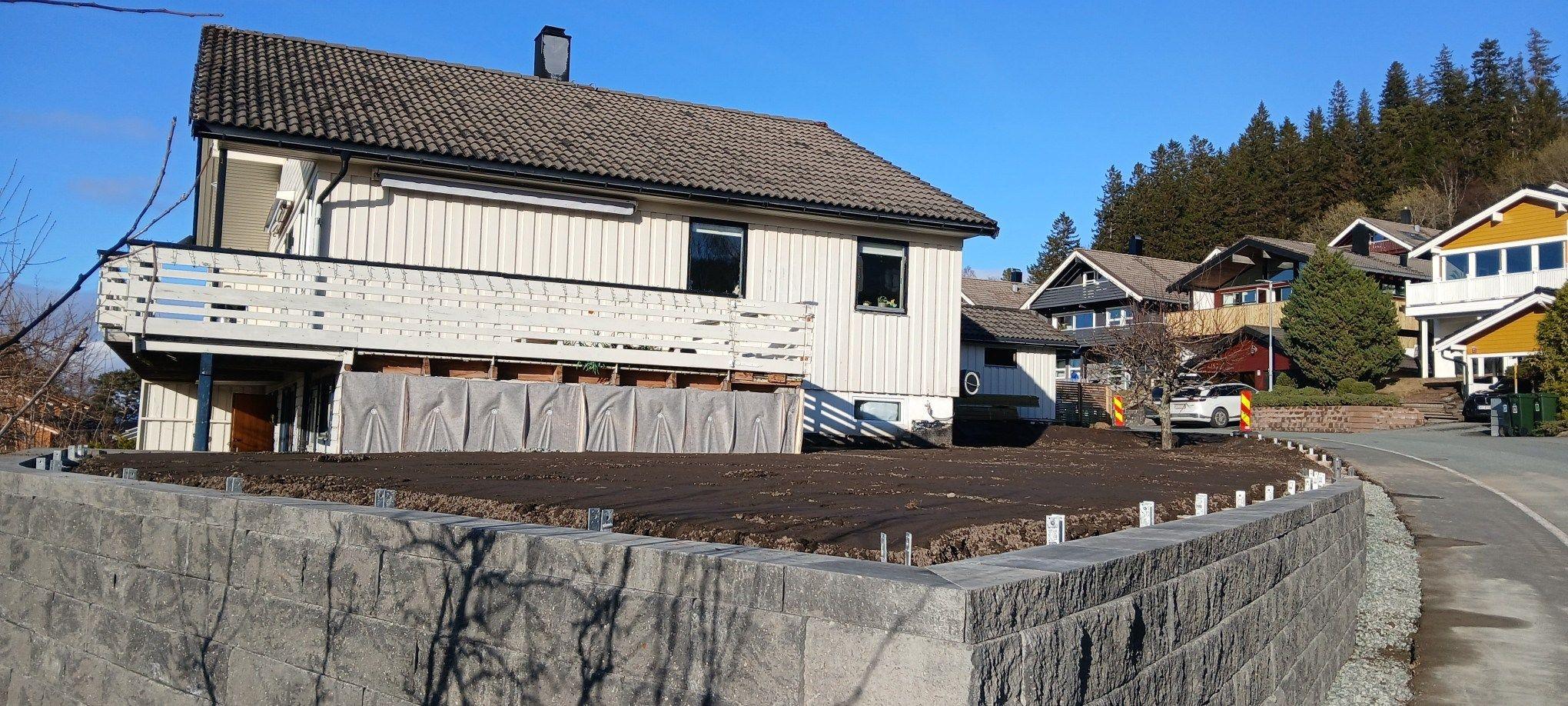 En hvit enebolig med terrasse, beliggende på en skråning, omgitt av trær og andre hus.