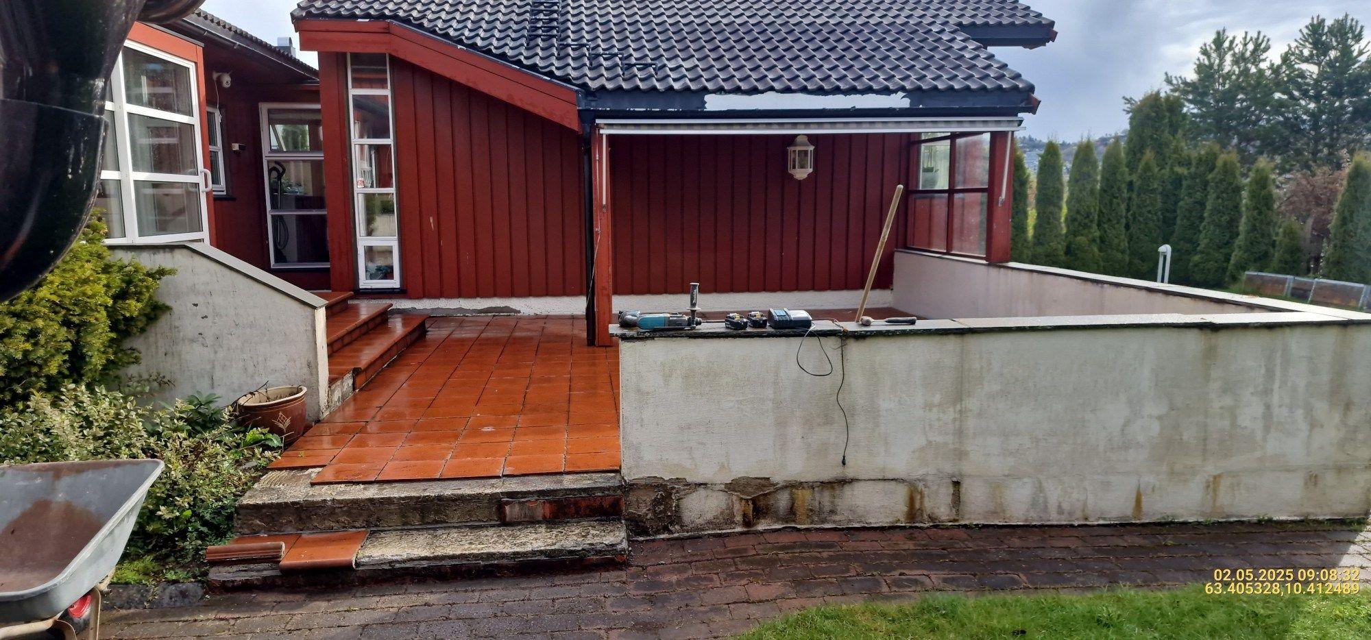 Hus med rød fasade, flislagt terrasse og hage, omkranset av grønne busker og trær.