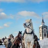 Hvit hest foran en kirke med snødekte bakker og ryttere i tradisjonelle klær i bakgrunnen.
