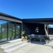 Moderne hus med store vinduer, terrasse og utemøbler under klar blå himmel.