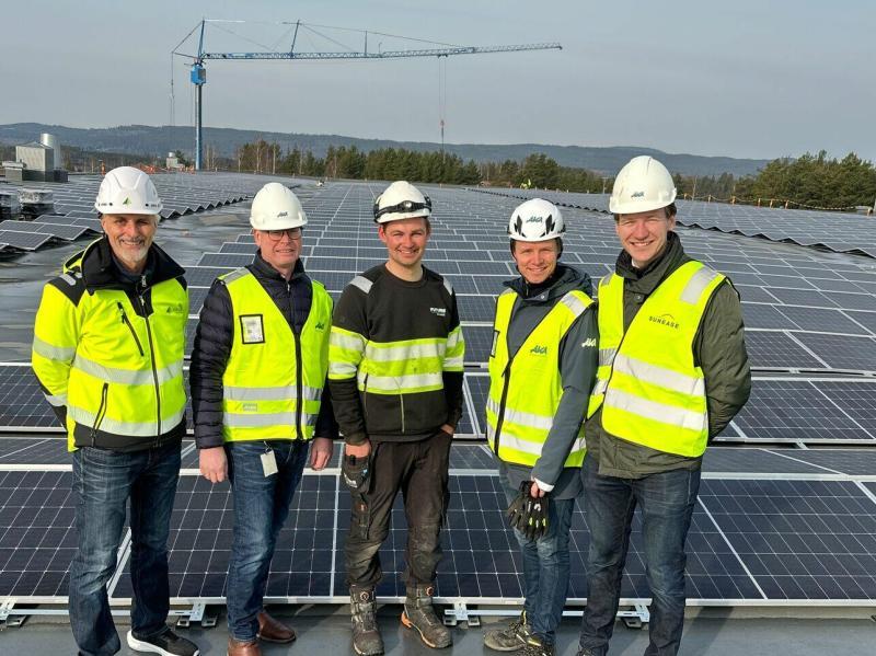 Fem menn i sikkerhetsutstyr står på et tak dekket med solcellepaneler, med en kran i bakgrunnen.
