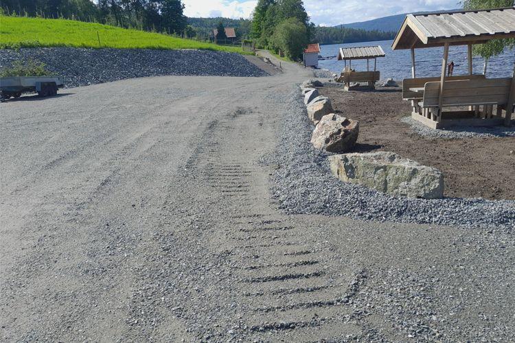 Grusvei ved vannet med spor etter kjøretøy, omgitt av steiner og benker under tak. Grønne områder i bakgrunnen.
