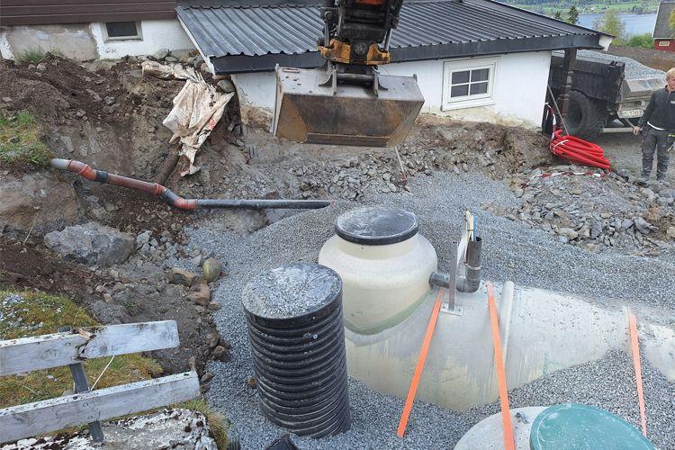 Gravearbeid med gravemaskin ved hus, med rør og septiktank synlig på grusunderlag.