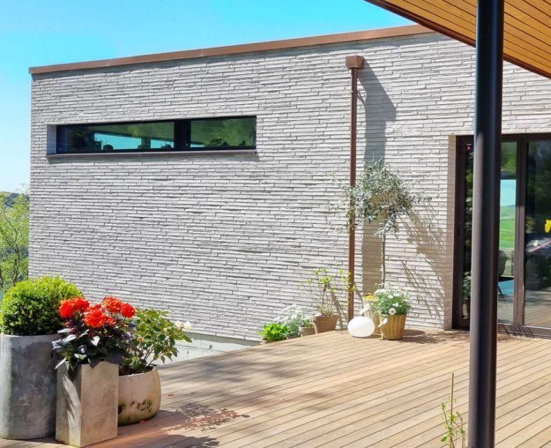 Moderne hus med steinvegg, store vinduer og blomsterpotter på terrassen. Solfylt dag med klar blå himmel.
