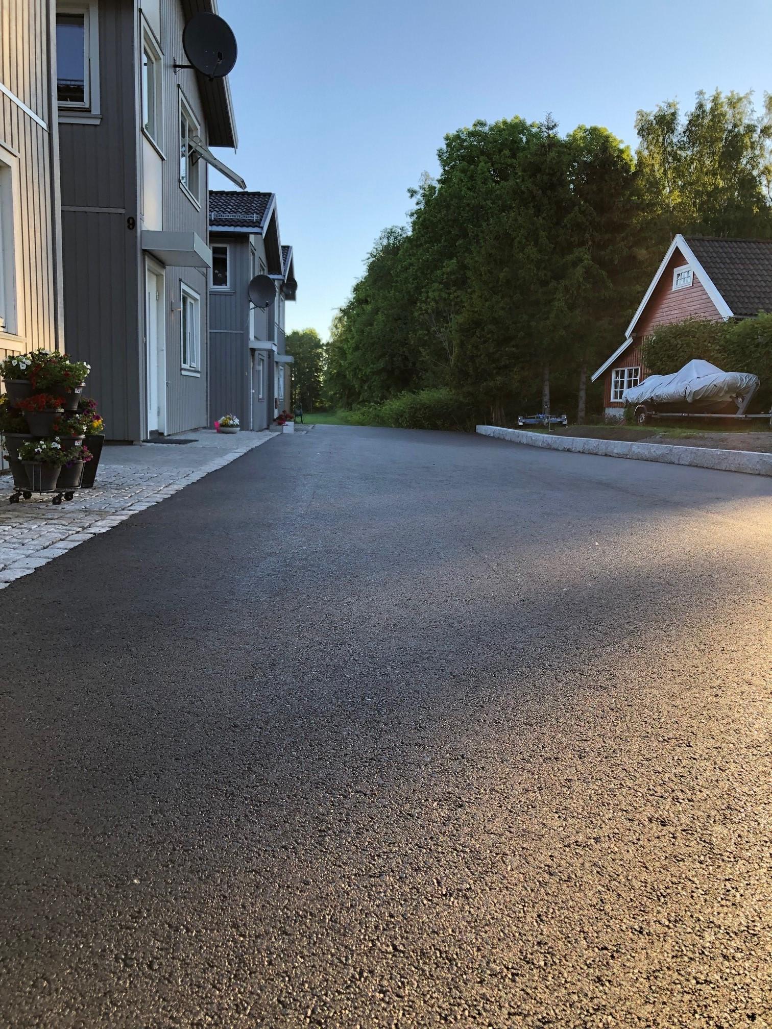 Ny asfaltlagt vei mellom hus med grøntområde og sollys i bakgrunnen.