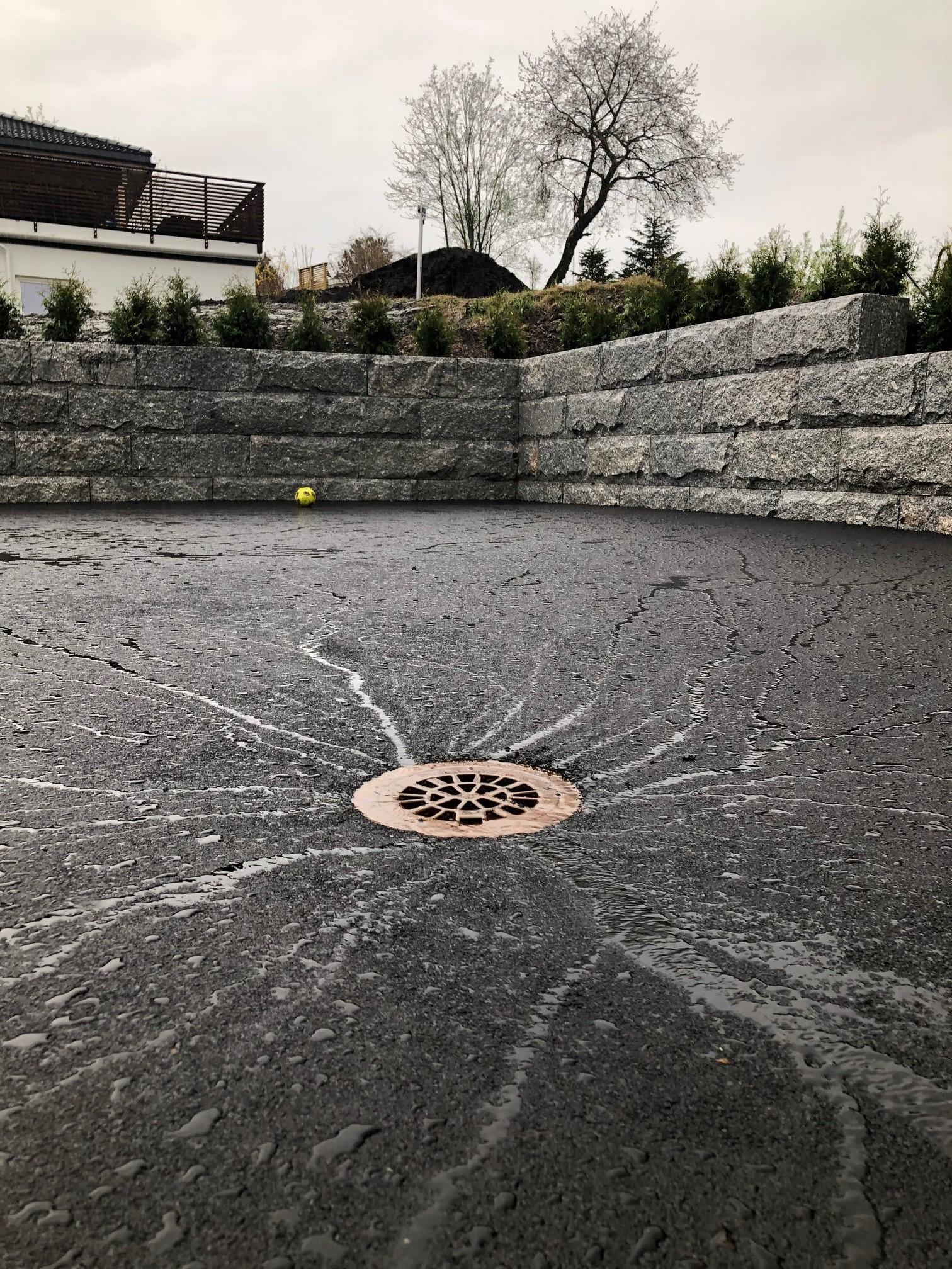 En grå betongoverflate med en avløpsrist i midten, omringet av steinmurer og grønne busker.