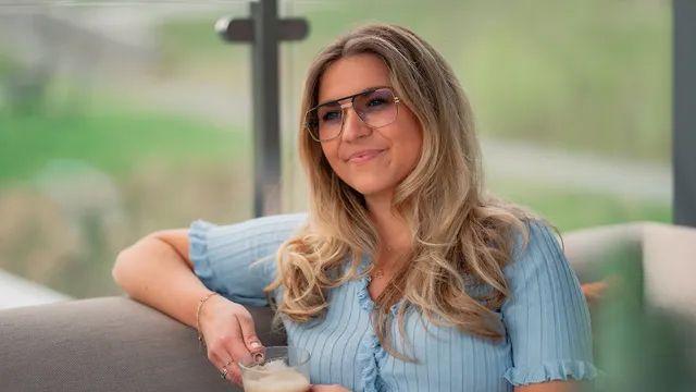 En kvinne med langt, bølgete hår sitter på en sofa og smiler mens hun holder en kopp. Grønne omgivelser i bakgrunnen.