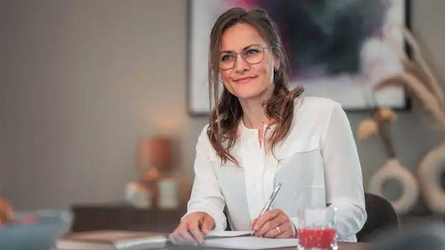 Kvinne med briller smiler mens hun skriver i notatbok, med moderne interiør i bakgrunnen.