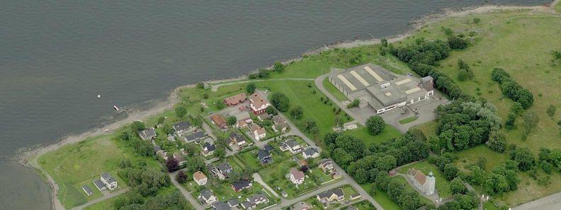 Oversiktsbilde av en kystby med hus, grøntområder og en fabrikk ved vannet.