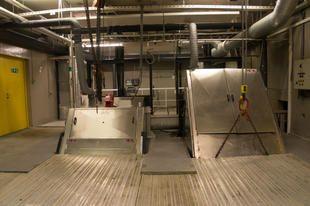 En industriell innendørs plass med to heiser og gule dører, omgitt av rør og elektriske paneler.