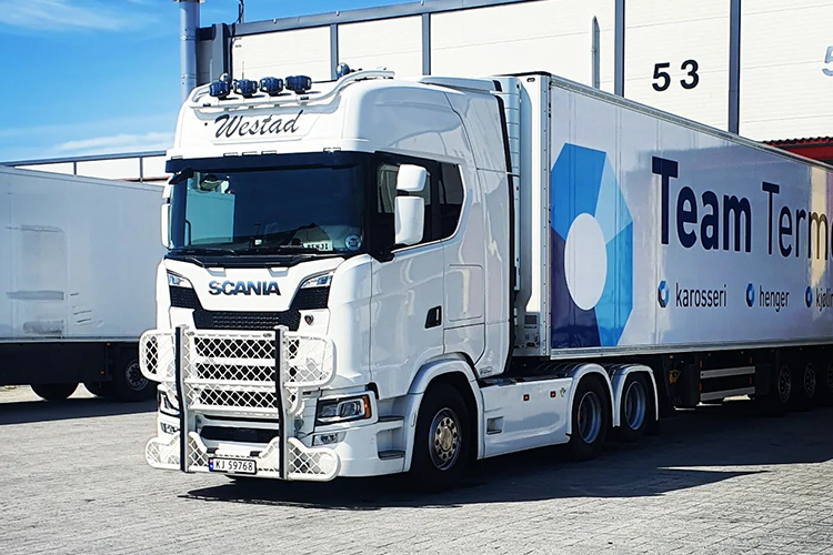En hvit Scania lastebil med tilhenger, merket "Team Termo", parkert ved en lagerbygning.