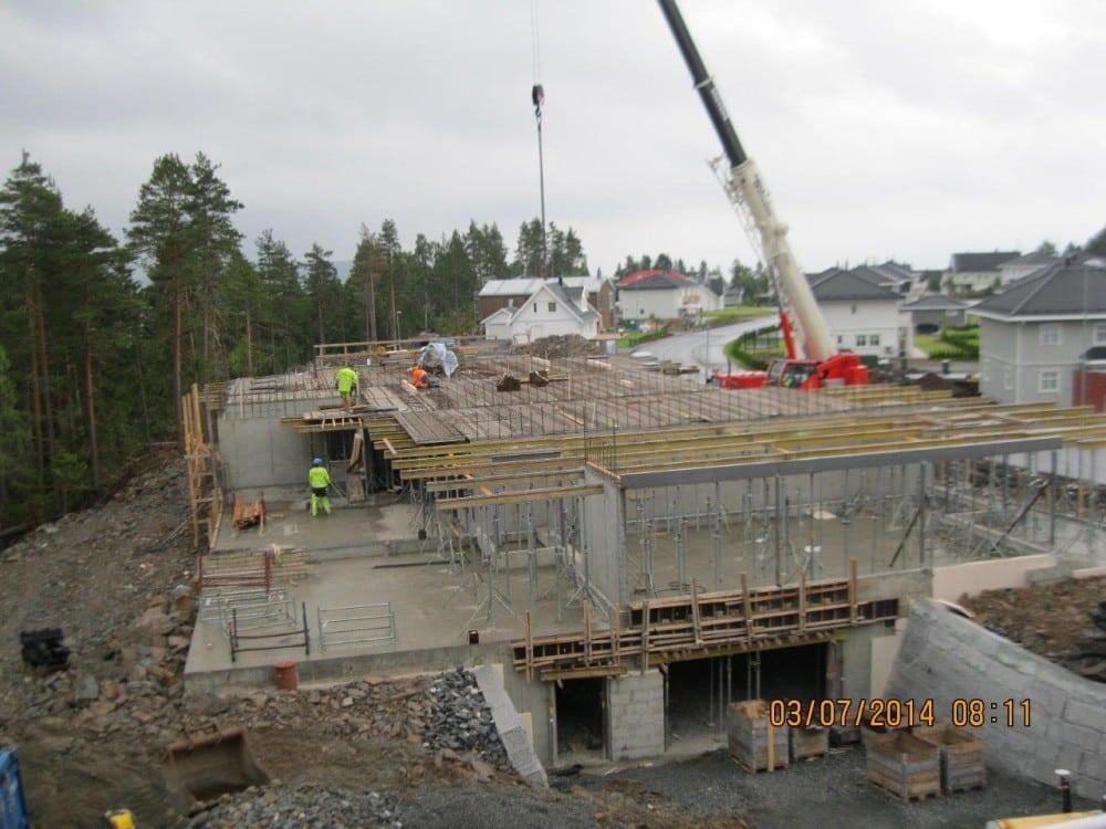 Byggeplass med arbeidere som jobber på en betongkonstruksjon under oppføring, omgitt av trær og boliger.
