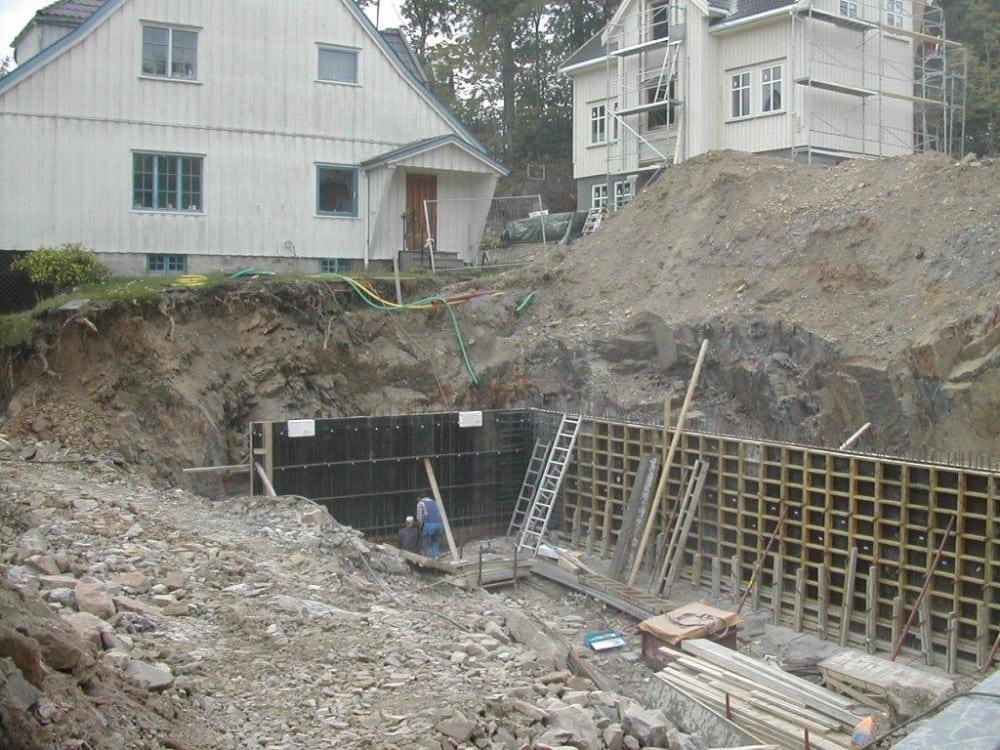 Byggeplass med utgravd grunn, forskaling og to hus i bakgrunnen. Arbeider i grøften.
