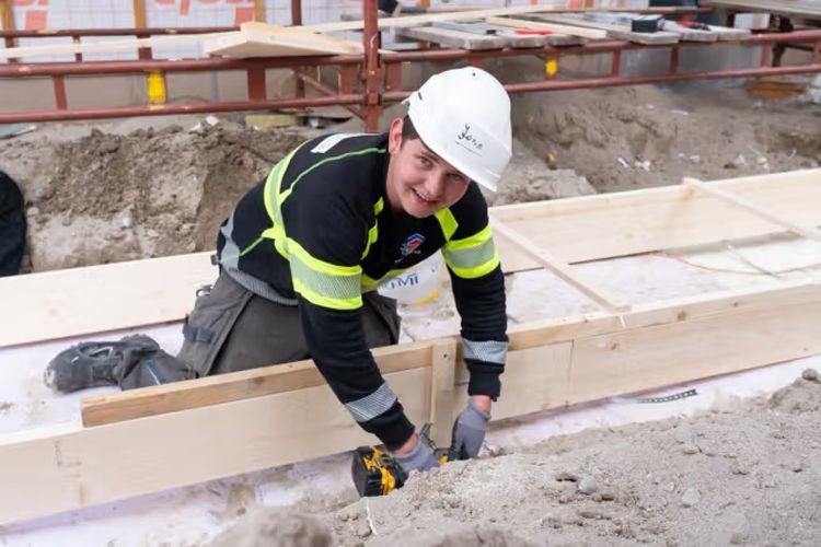 En håndverker i hjelm og arbeidsklær jobber med treverk på en byggeplass.