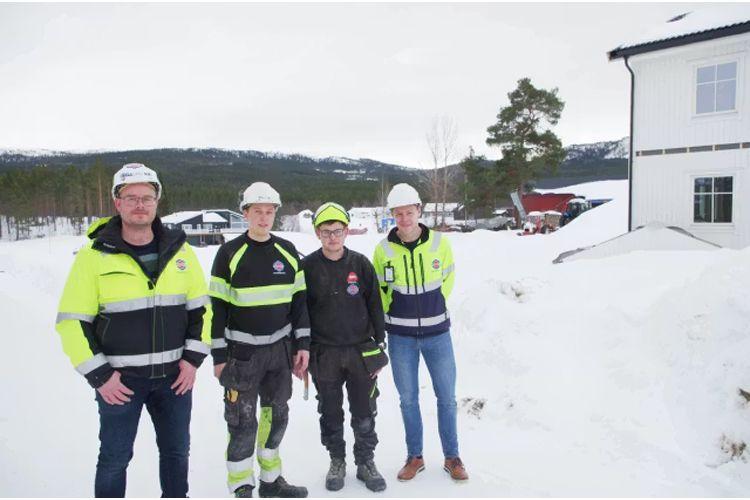 Tre menn i arbeidsklær og hjelmer står i snøen foran en bygning, med fjell i bakgrunnen.
