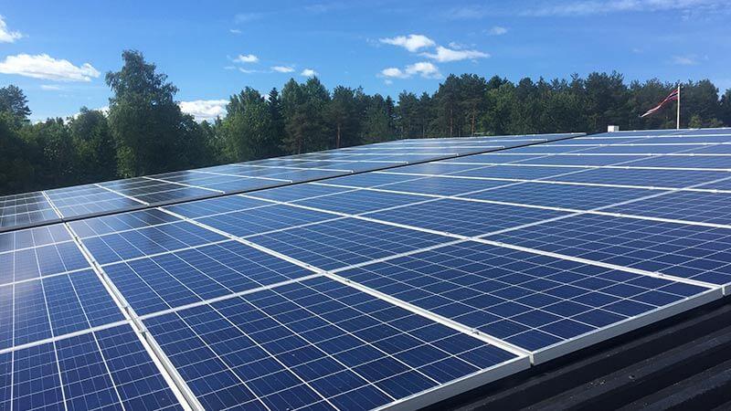 Solcellepaneler på taket med grønn skog i bakgrunnen og blå himmel med skyer.