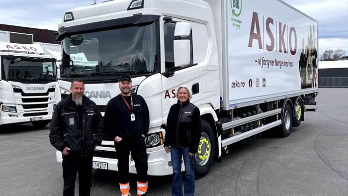 Tre personer står foran en Scania lastebil med ASKO-logo, på en parkeringsplass.