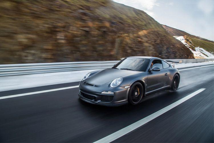 En grå Porsche 911 kjører raskt på en vei med snødekte fjell i bakgrunnen.