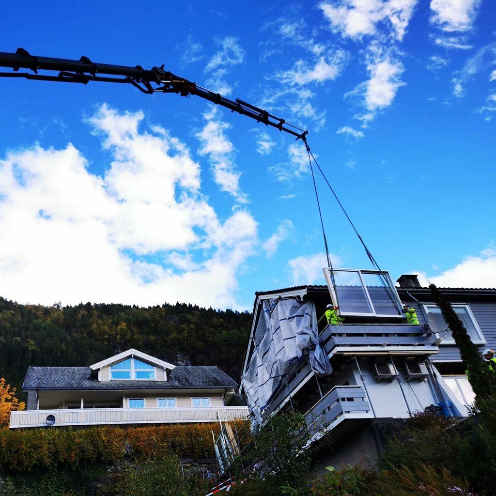Kran løfter en balkong fra en bygning under en klar blå himmel med skyer.