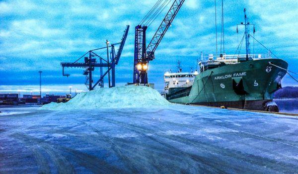 Lasteskip ved kai med kraner og en stor haug med salt i havnen under en blå himmel.