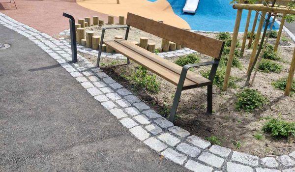 Benk i en park med steinlagt sti og lekeområde i bakgrunnen, omgitt av grønne planter.