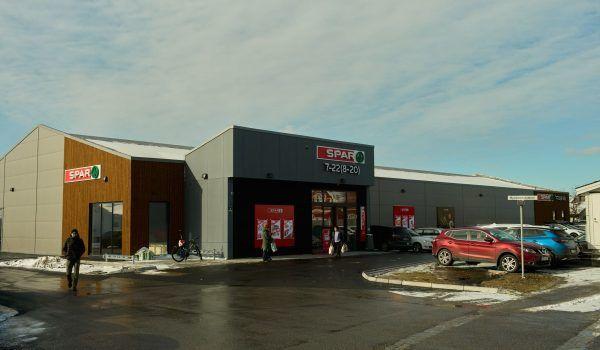 En moderne butikkbygning med store vinduer, parkerte biler og snø på bakken.