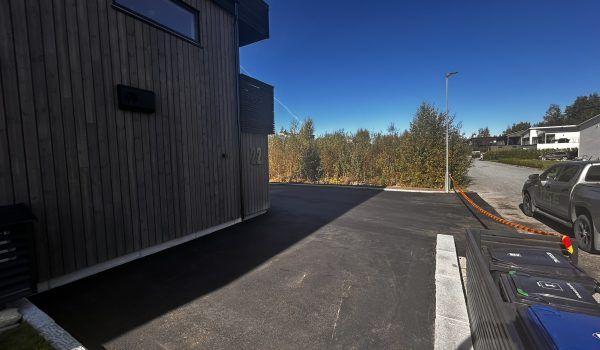 Moderne bygning med trepanel og asfaltert parkeringsplass under klar himmel. Grønt område i bakgrunnen.