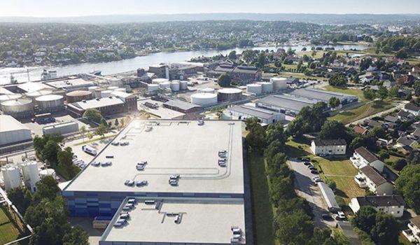 Oversiktsbilde av et industriområde med bygninger, lagertanker og elv i bakgrunnen. Solfylt dag.
