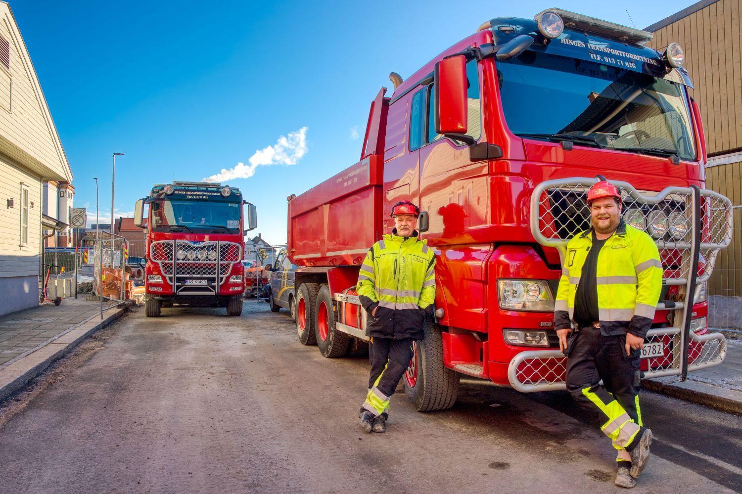 To personer i arbeidsklær står ved siden av to røde lastebiler på en gate med blå himmel i bakgrunnen.