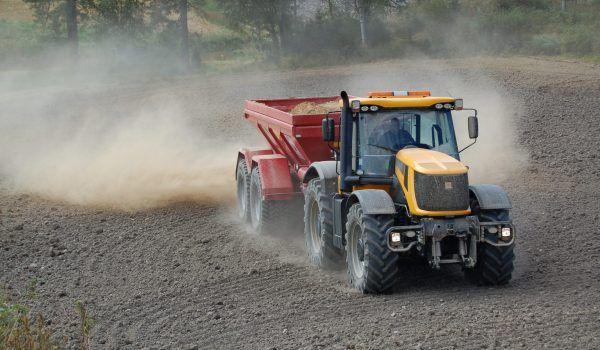 En traktor som kjører på en åker og drar en rød tilhenger med jord, omringet av støv.