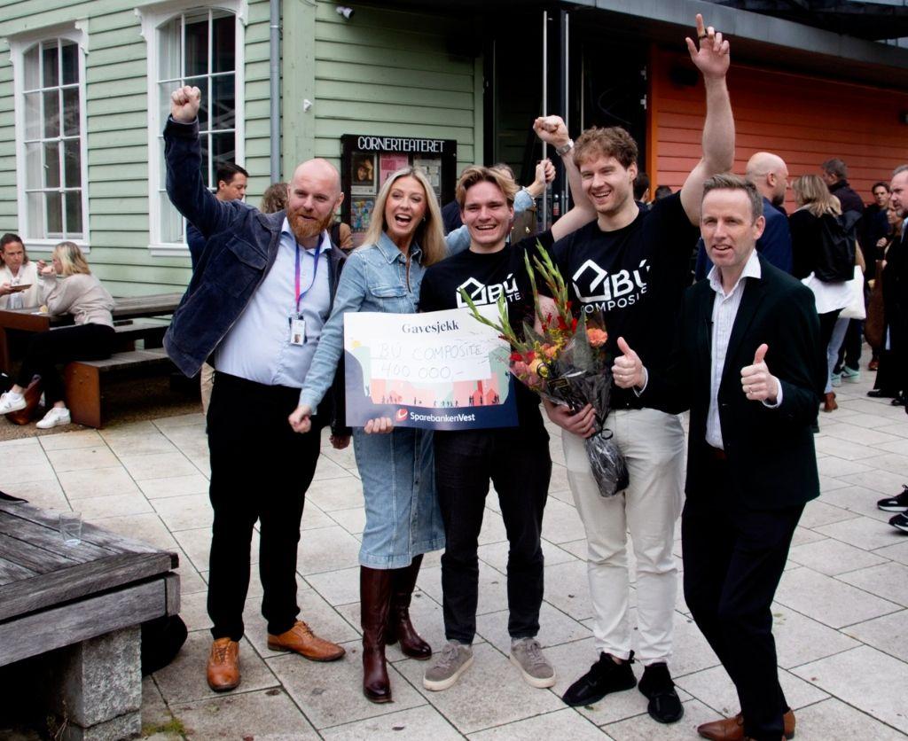 Gruppe mennesker feirer med blomster og plakett utenfor en bygning, alle smilende og hever hendene i jubel.